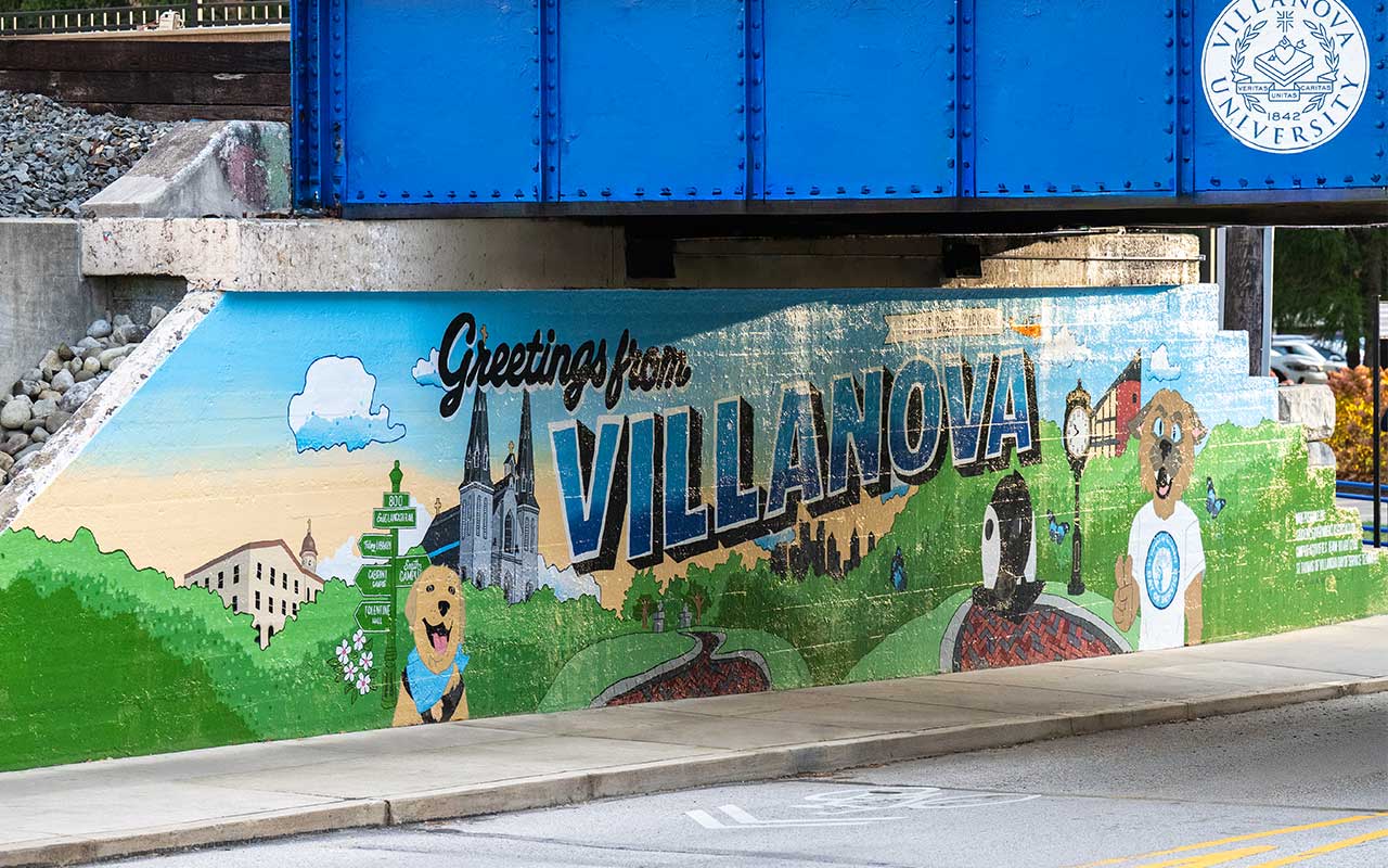 A colorful mural under a bridge reads “Greetings from Villanova” and features illustrated campus landmarks, a church, street signs, and Villanova mascot imagery.