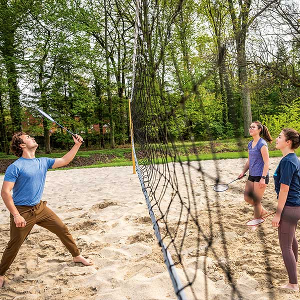 People play badminton on a sand court separated by a net, surrounded by trees.