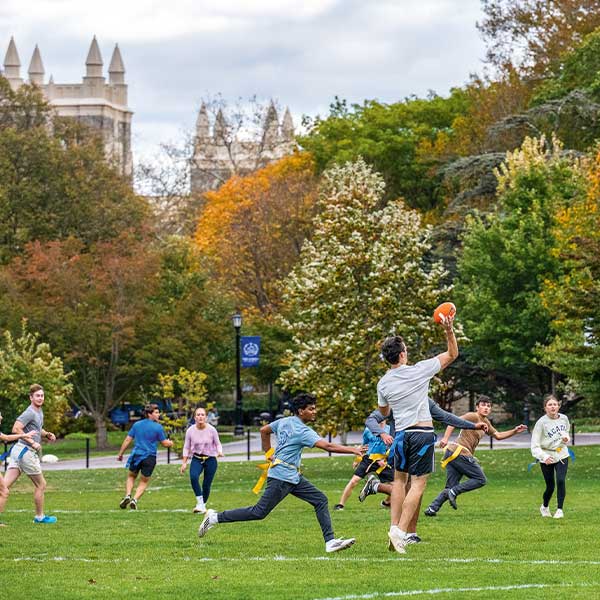 A group of people play flag football on a grassy field with campus buildings in the background.