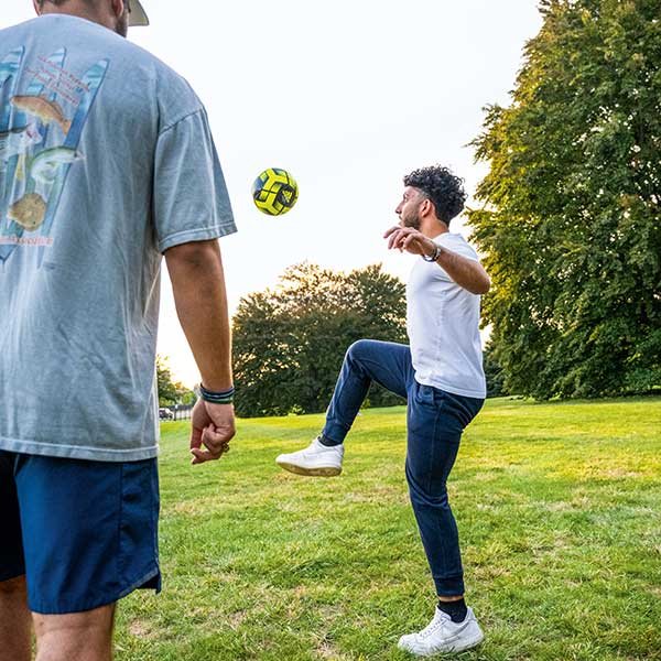 Two people stand on a grassy lawn as one kicks a soccer ball into the air.