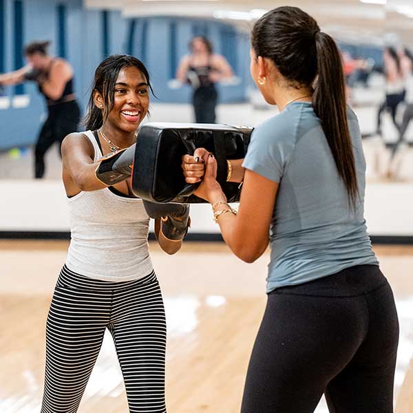 Two people face each other in a fitness studio while using padded training equipment.