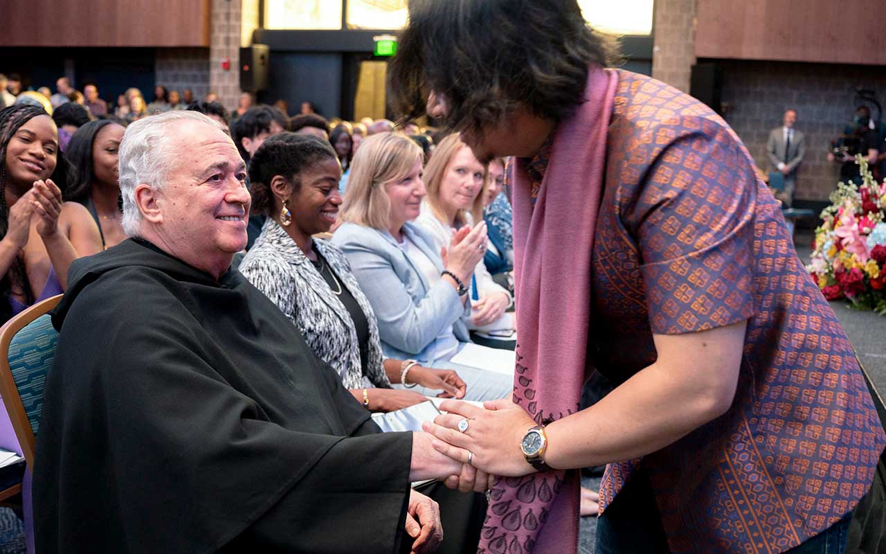 Father Peter sits among an audience while greeting an attendee who leans forward to shake his hand.