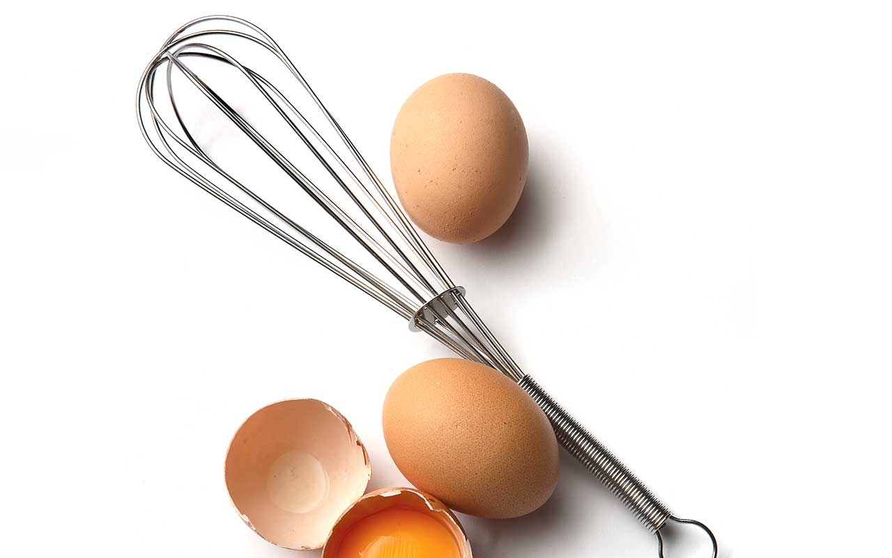 A metal whisk rests beside whole eggs and cracked eggshells on a white background.
