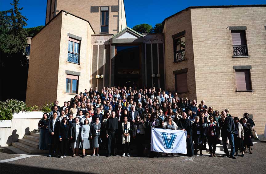 Large group of Villanova University students, faculty and alumni gathered on the steps outside the Augustinianum building, with a Villanova “V” flag displayed at the front