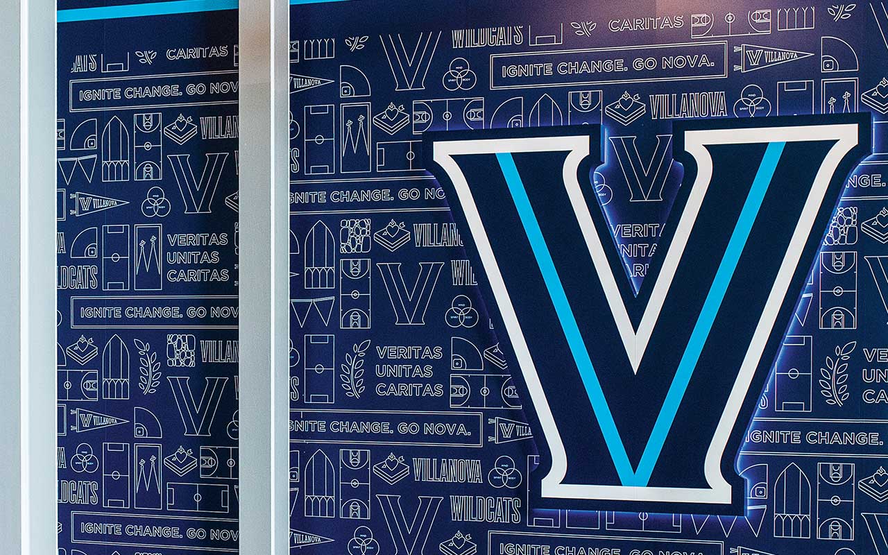 A large illuminated blue “V” logo is mounted inside a building against a patterned wall featuring Villanova words, symbols, and athletic iconography.