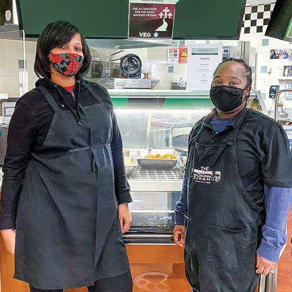 two women wearing aprons stand in front of a food case